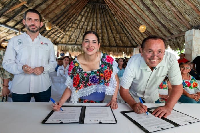 Tres personas firman documentos en una mesa bajo una estructura con techo de palma, con asistentes de fondo en un evento comunitario.