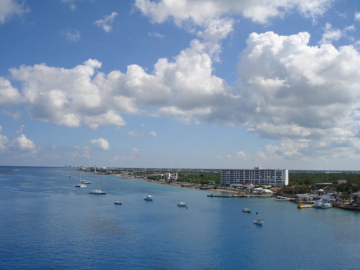 Vista aérea de una bahía costera turquesa con yates y lanchas fondeadas frente a un hotel blanco y edificaciones de un centro turístico bajo un cielo con nubes cúmulos.
