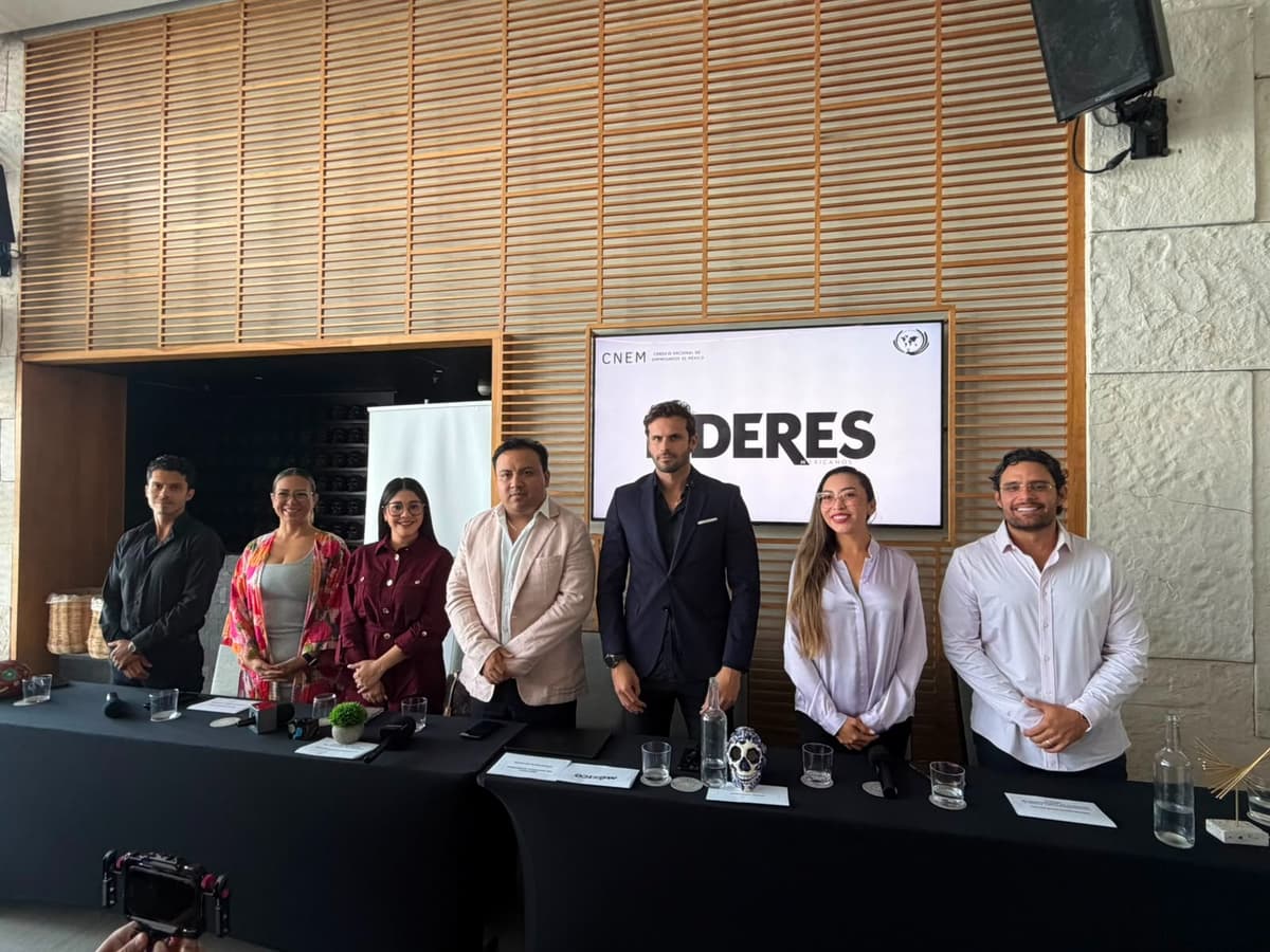 Siete personas en traje de negocios de pie frente a una mesa con micrófono durante una conferencia, con pantalla de fondo que muestra texto sobre emprendimiento.