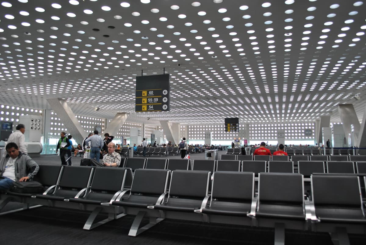 Sala de espera de una terminal aeroportuaria con asientos metálicos vacíos, pasajeros esperando y letreros que indican las puertas 54 a 60, bajo un techo perforado de luces.